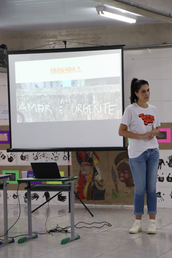 Mulher com camiseta da turma do jiló ao lado de um projetor com os dizeres "Amar é Urgente".