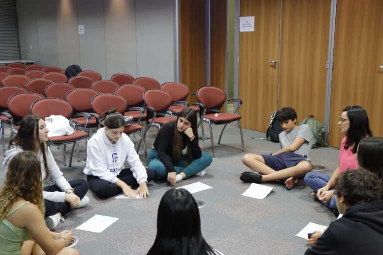Jovens sentados em círculo no chão em um auditório escolar.